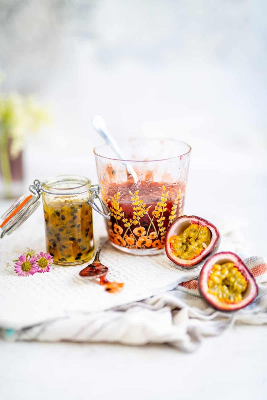 A glass jar of Passion Fruit Jam, a bowl of red fruit sauce with a spoon, and two halved passionfruits are arranged on a white cloth with a patterned towel and small flowers nearby.