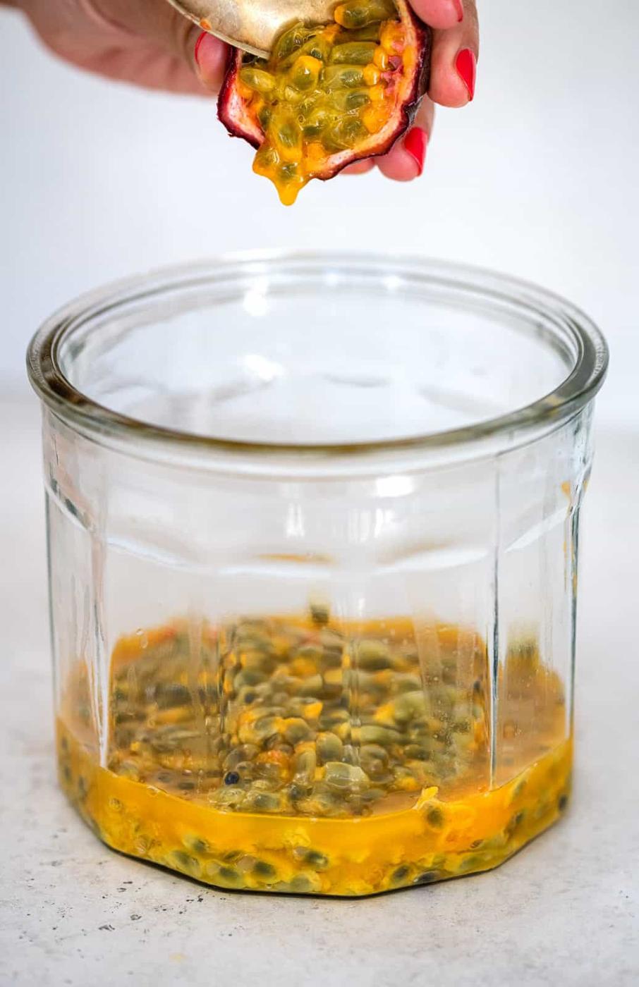 A hand squeezes half a passion fruit, pouring its yellow pulp and seeds into a clear glass jar on a light surface—perfect for making homemade passion fruit jam.