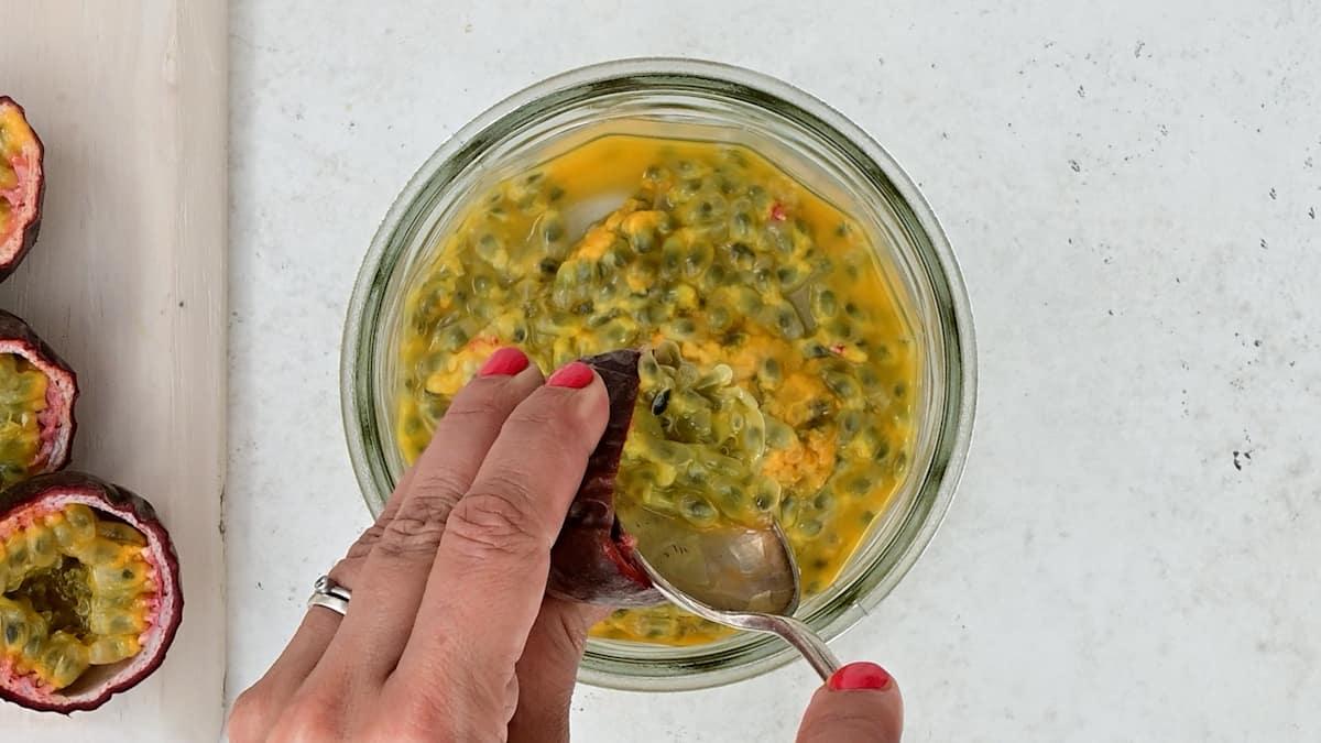 A hand holding a spoon scoops passion fruit pulp from a halved passion fruit into a glass jar of homemade Passion Fruit Jam, on a light-colored surface.