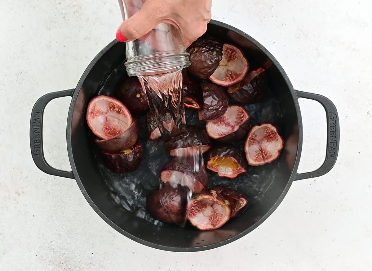 A hand pours water from a glass jar into a black pot filled with halved passion fruits on a white surface, preparing to make Passion Fruit Jam.