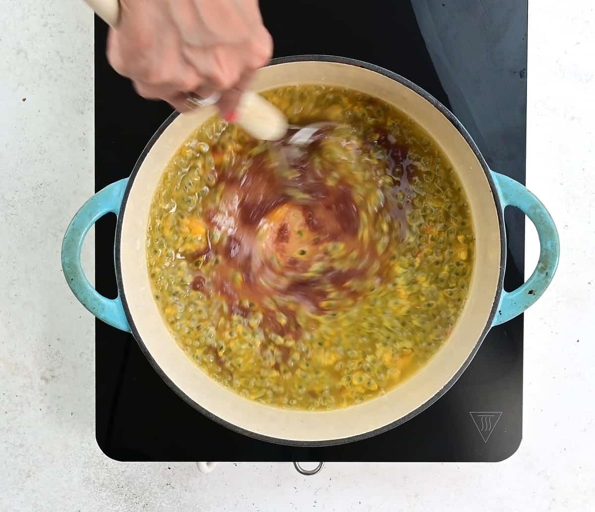 A hand stirs passion fruit jam, its yellow and green hues swirling together with a wooden spoon in a blue-handled pot on a black stovetop.