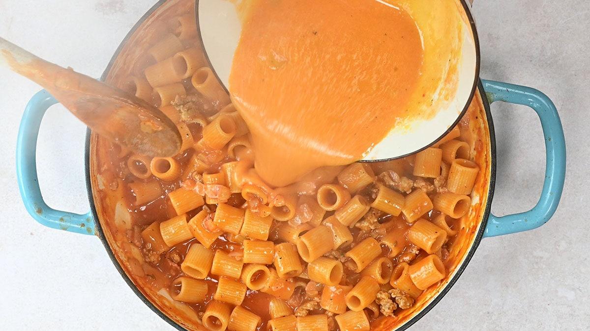 A blue pot filled with rigatoni pasta and ground sausage is being topped with tempered egg yolk and cheese mixture poured from a white dish. A wooden spoon rests in the pot, and steam rises from the hot ingredients.