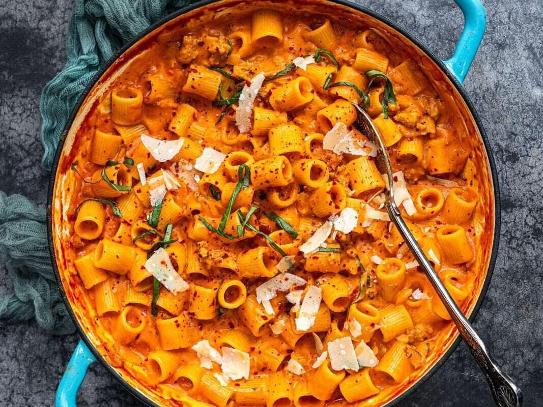 A turquoise pot filled with creamy tomato-basil pasta garnished with Parmesan cheese and fresh basil sits on a textured gray surface. A silver serving spoon is placed inside the pot, next to a loosely draped green cloth.