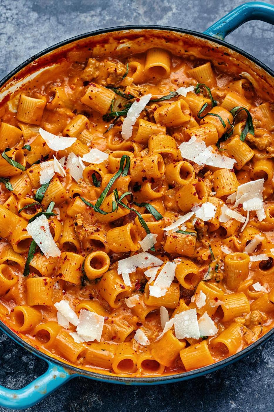 A pan filled with creamy rigatoni pasta in a rich red sauce, topped with shaved parmesan cheese and garnished with fresh herbs. The pasta is cooked al dente, and the vibrant sauce is speckled with spices, creating an inviting, hearty dish.