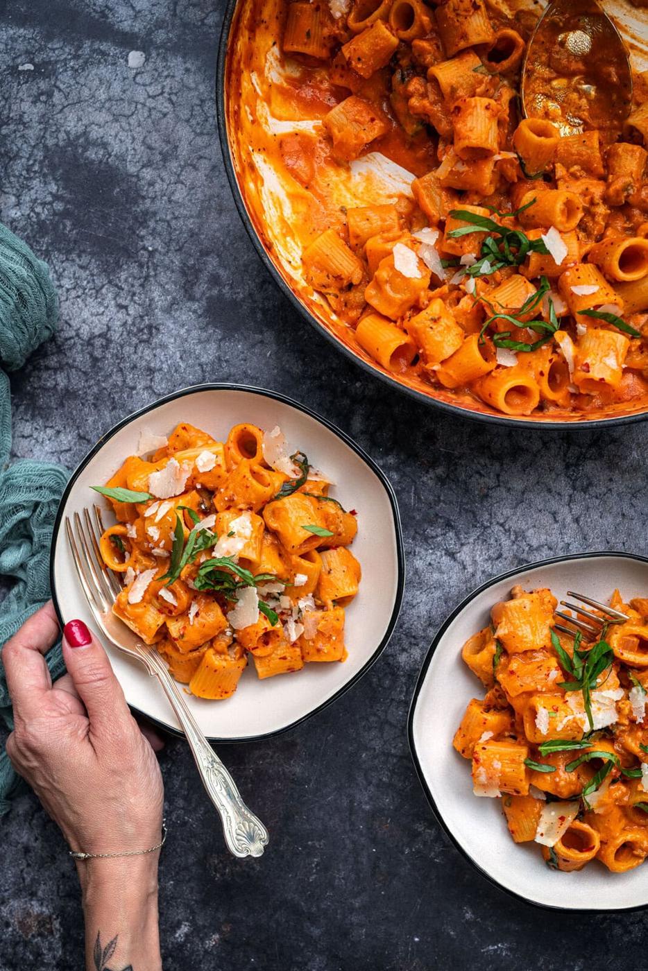 Close-up of creamy rigatoni pasta served in two bowls, garnished with herbs and cheese. One hand holds a fork and touches a bowl. A pan with more pasta is visible in the background. The scene is set on a dark textured surface.