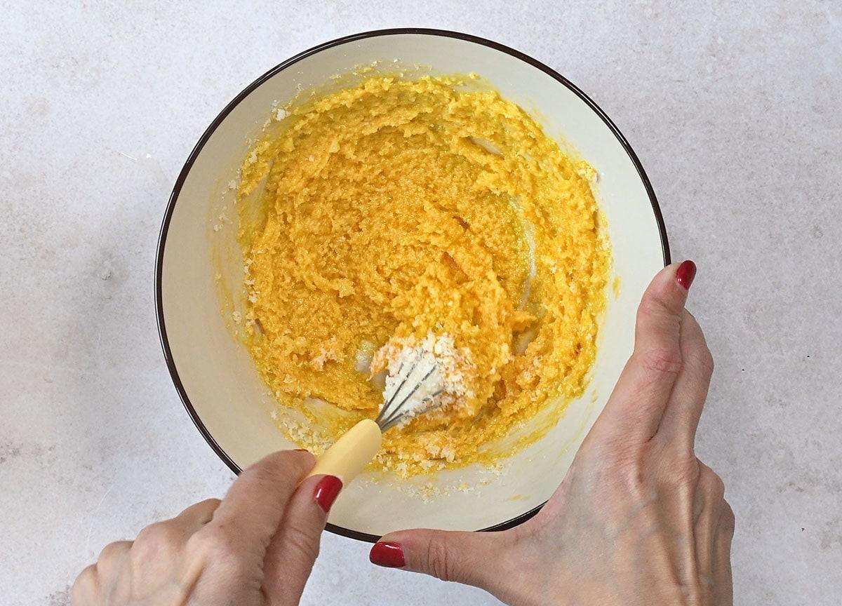 Hands with red nail polish mixing a egg yolks and finely grated cheese in a white bowl using a fork. The bowl is on a light-colored surface.