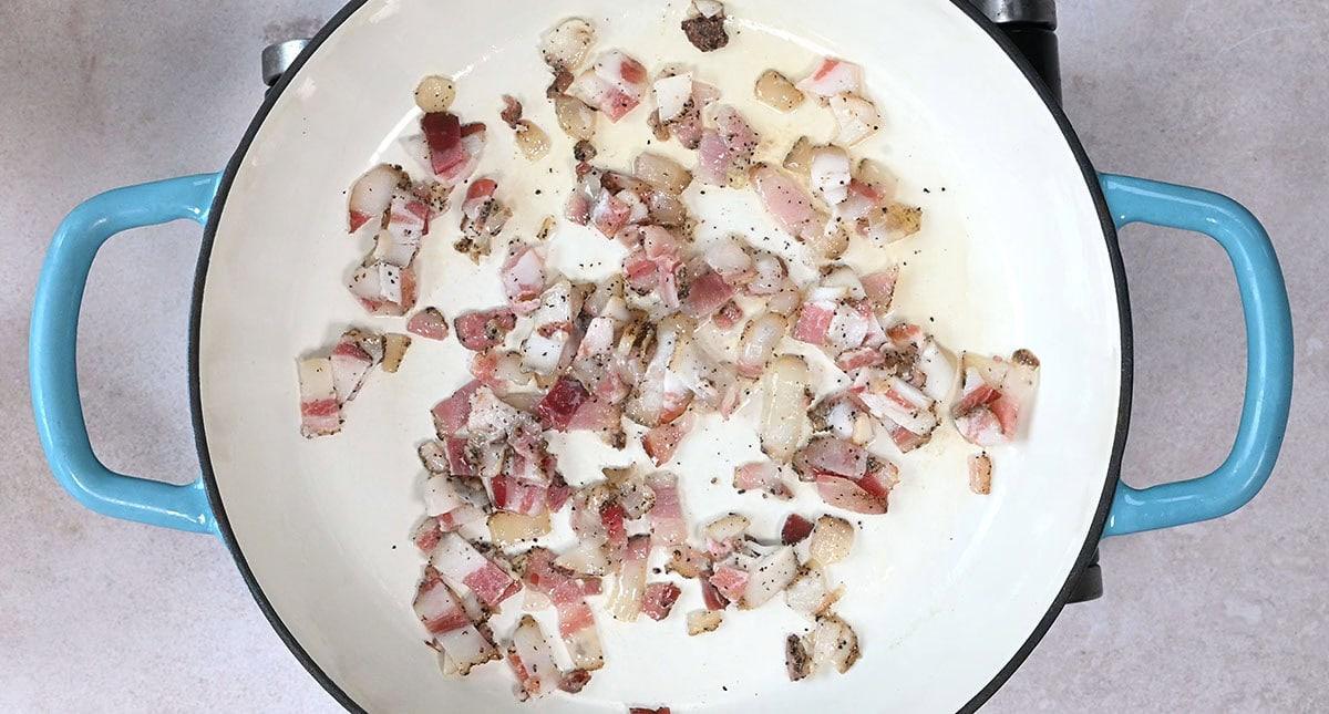 Chopped guanciale pieces cooking in a light-colored skillet with blue handles. The bacon is sizzling and looks slightly browned, with a mix of fat and lean parts visible.