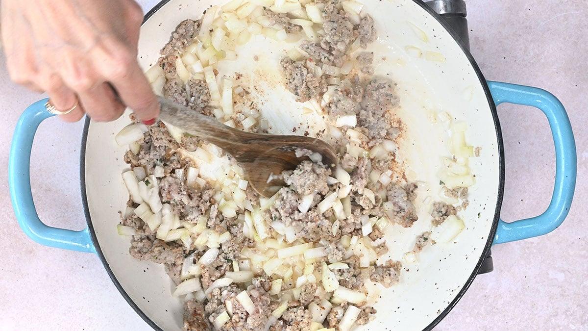 A hand stirs sausage meat and chopped onions in a large blue-handled skillet with a wooden spoon. The mixture is beginning to brown and cook, creating a savory dish.