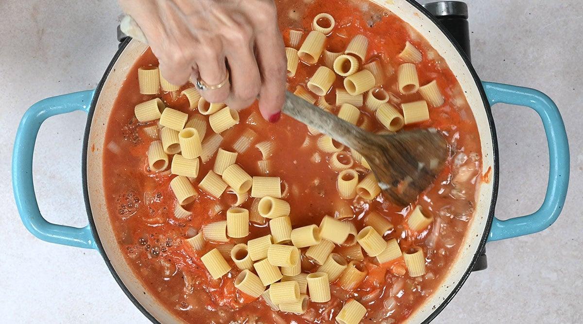 A person stirs pasta tubes into a pot of tomato sauce with a wooden spoon. The pot has blue handles and is set on a light countertop.