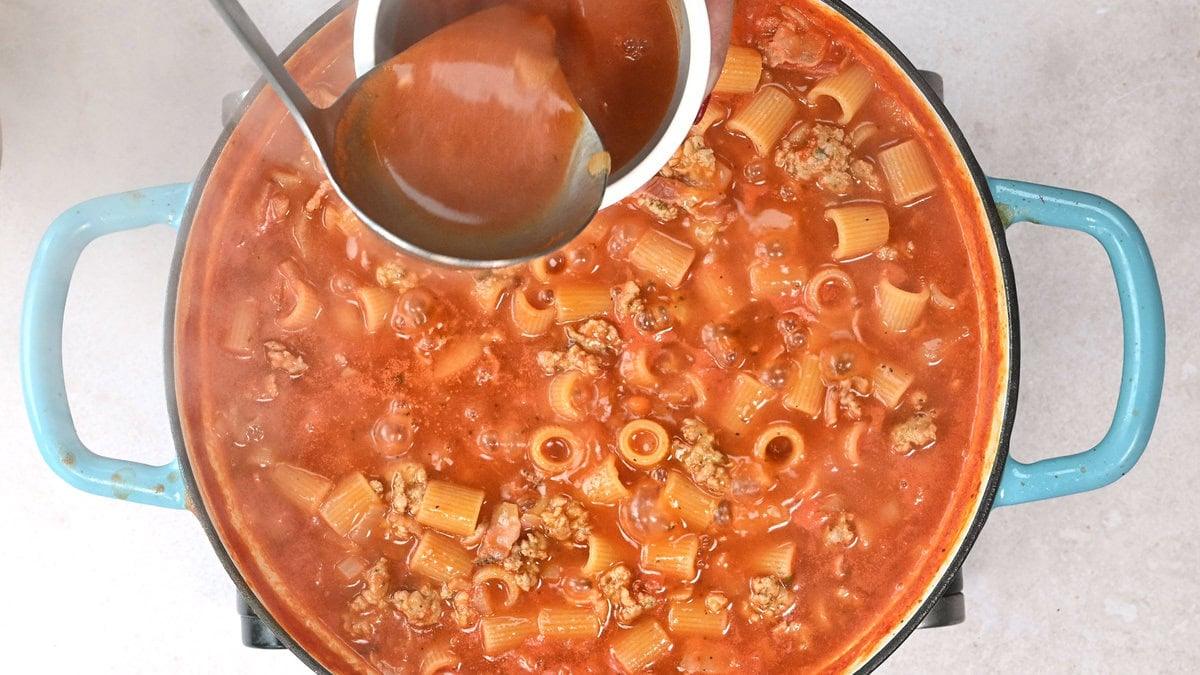 A pot of pasta soup with rigatoni, sausage, and tomato-based broth is simmering on a stove. A ladle is pouring additional broth into the pot. The pot has blue handles and is sitting on a light-colored surface.