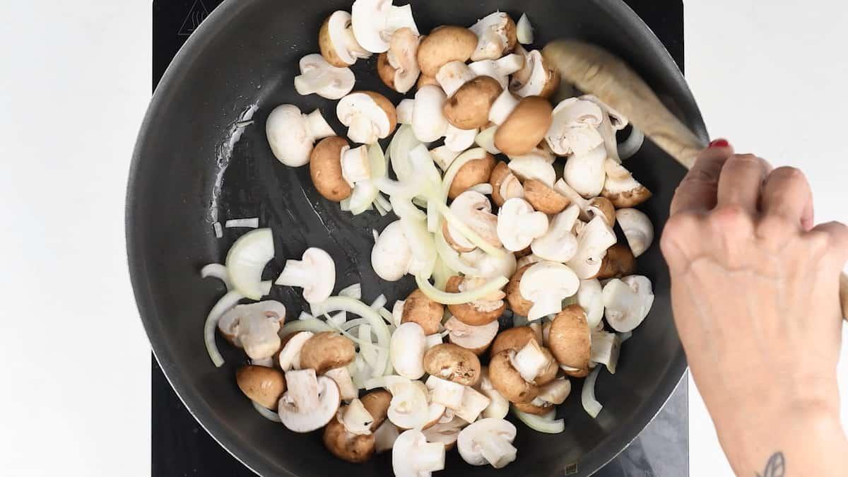 A person expertly stirs sliced mushrooms and onions in a black frying pan on the stove, preparing the savory base for a delicious pasta bake. Holding a wooden spatula, with a tattoo visible on their hand, they skillfully blend white and brown mushroom varieties.