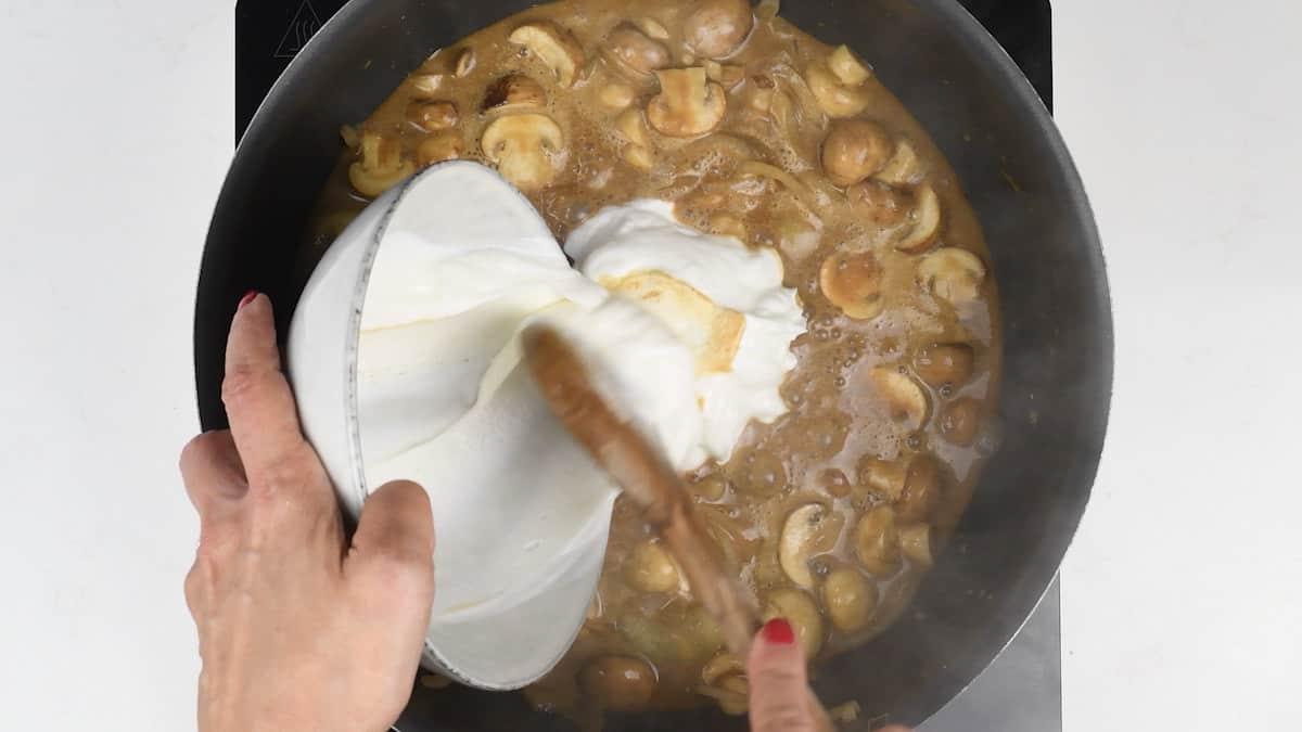 A person with red nail polish is pouring a creamy white sauce into a skillet filled with mushrooms, stirring it with a wooden spoon. The scene on the stovetop hints at the start of a delightful pasta bake, all set against an inviting white backdrop.
