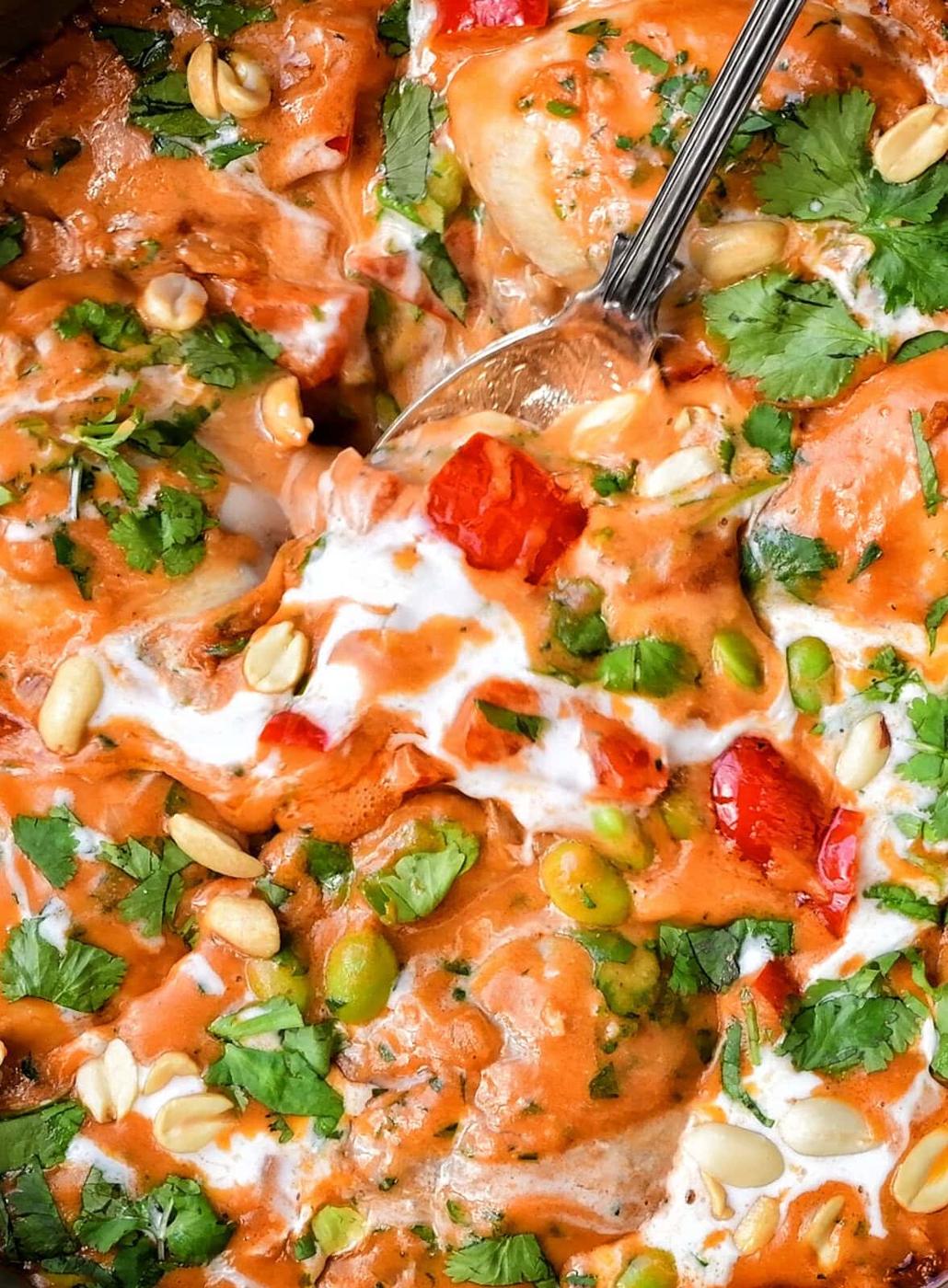 A close-up of a creamy, peanut butter chicken dish topped with chopped cilantro, peanuts, edamame, and red bell peppers, with a spoon partially submerged in the food. Swirls of coconut cream are visible on the surface.