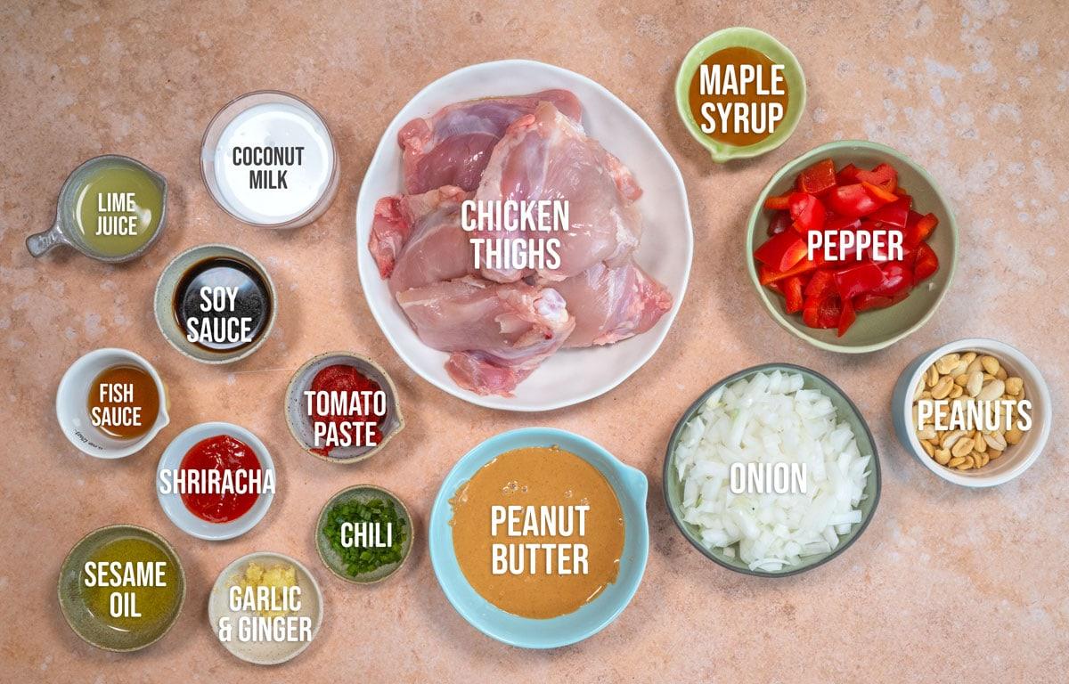 A top-down view of a table with labeled ingredients for a recipe, including chicken thighs, peanut butter, soy sauce, coconut milk, red pepper, peanuts, onion, garlic & ginger, lime juice, sesame oil, fish sauce, sriracha, tomato paste, chili, and maple syrup.