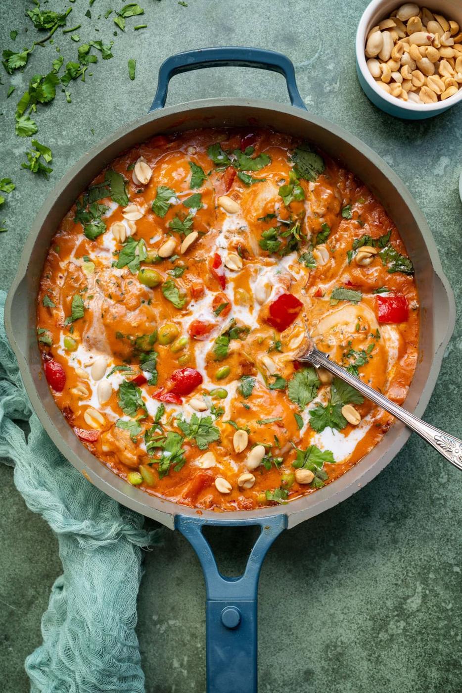 A pot of creamy peanut stew with chicken, red peppers, green peas, and cilantro, topped with peanuts and a swirl of cream, with a spoon inside. A small bowl of peanuts and chopped cilantro are nearby.
