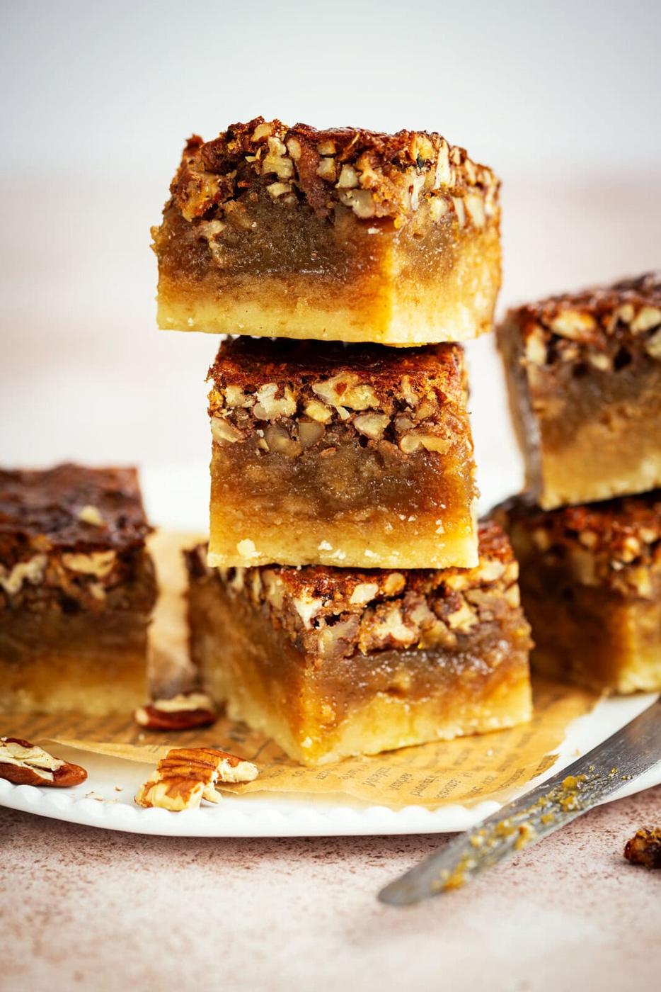 Three pecan pie bars stacked on a plate, with additional bars and pecan pieces around them. A knife lies nearby on the plate, showing the gooey filling and crumbly crust layers of the dessert bars.