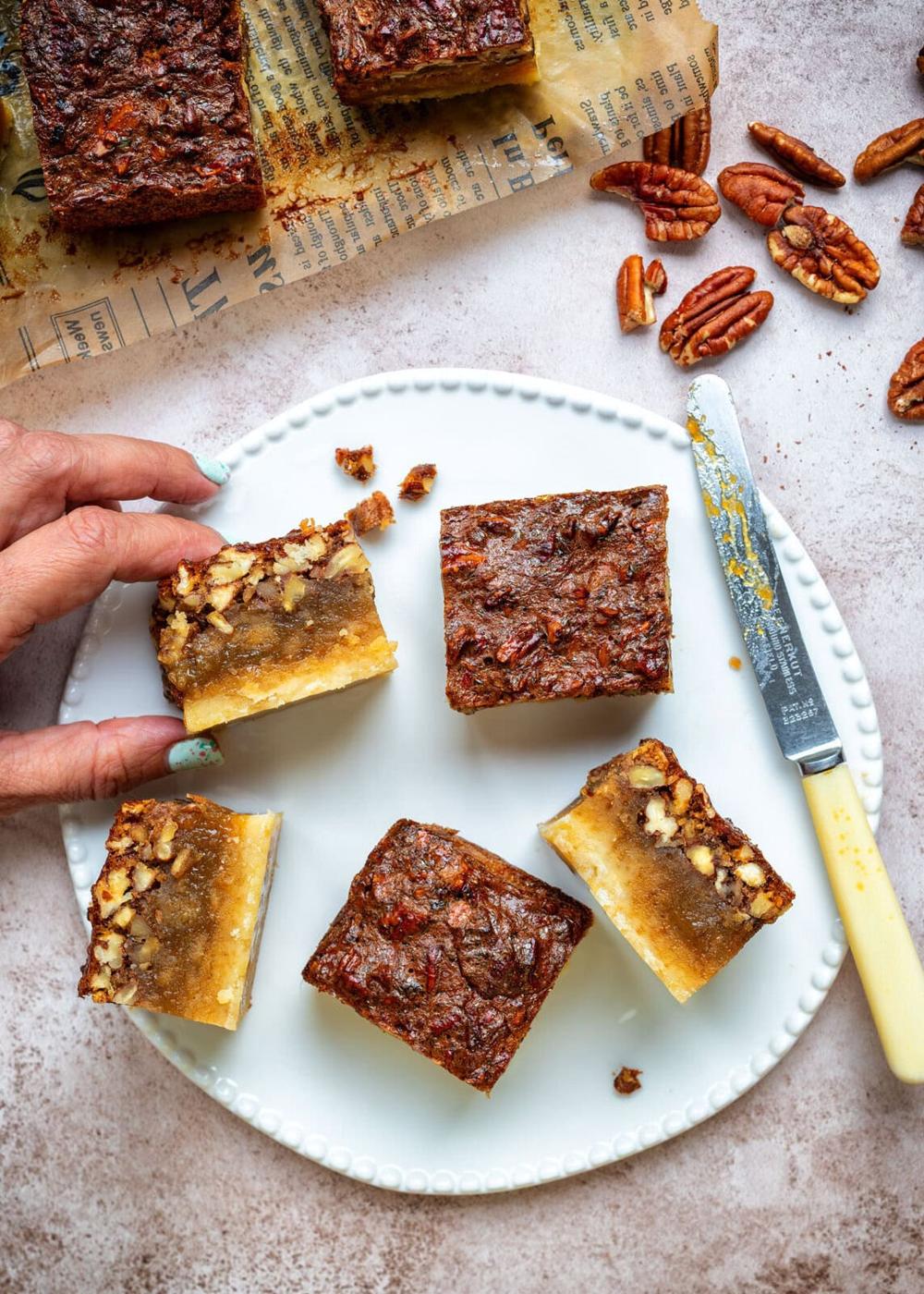 A hand reaches for a pecan pie bar on a white plate with five bars. A knife with a yellow handle, pecans, and more bars on brown paper are nearby on a light-colored surface.