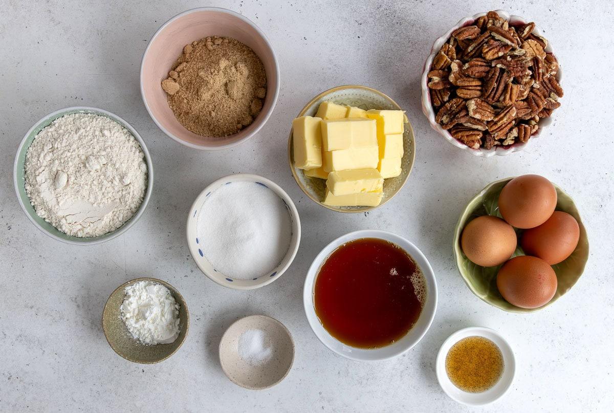 Bowl of flour, bowl of brown sugar, bowl of pecans, cubed butter, granulated sugar, eggs, syrup, cornstarch, salt, and vanilla extract arranged on a light surface.