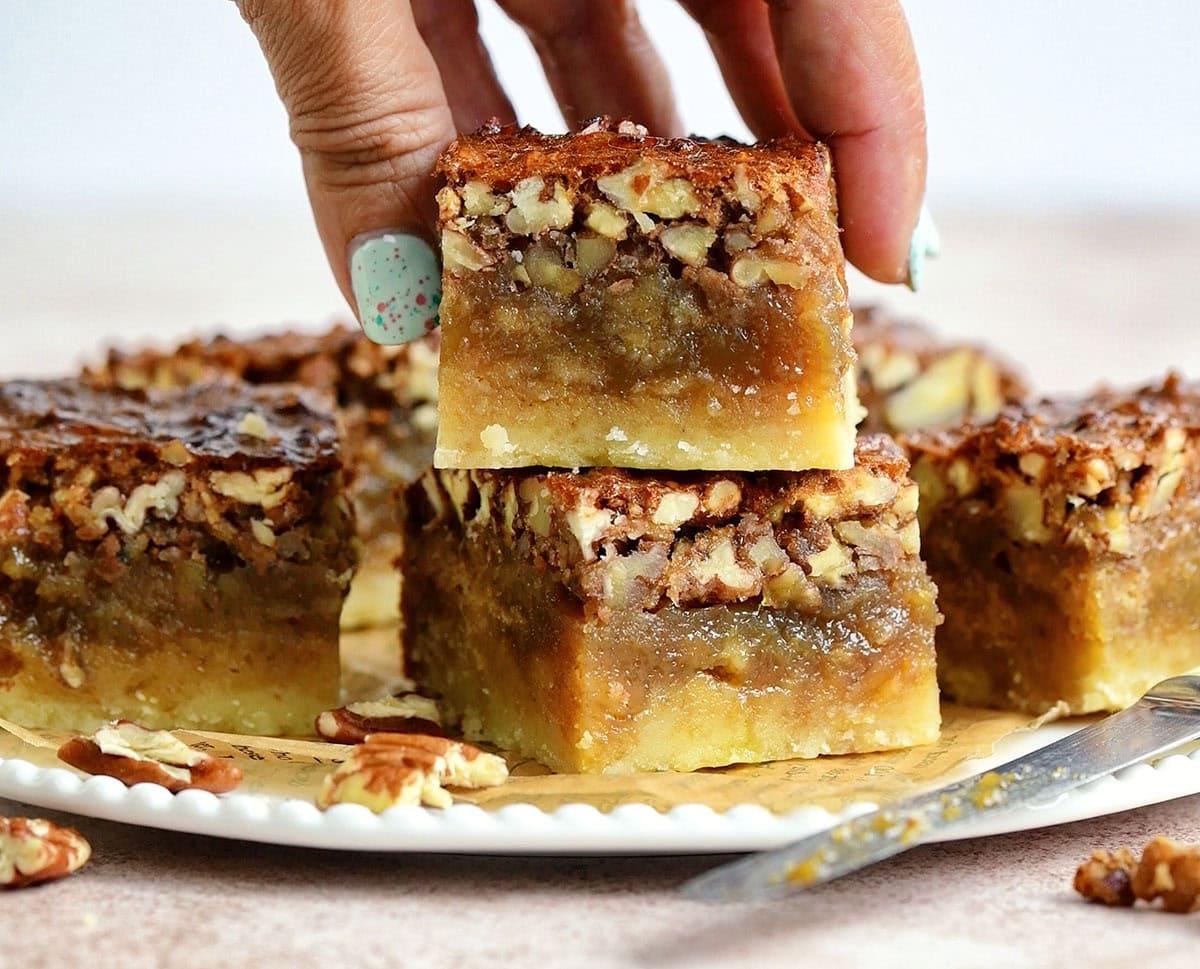 A hand with teal-painted nails picks up a pecan bar from a plate stacked with several gooey, nut-topped bars. A butter knife and scattered pecans are also visible on the plate.