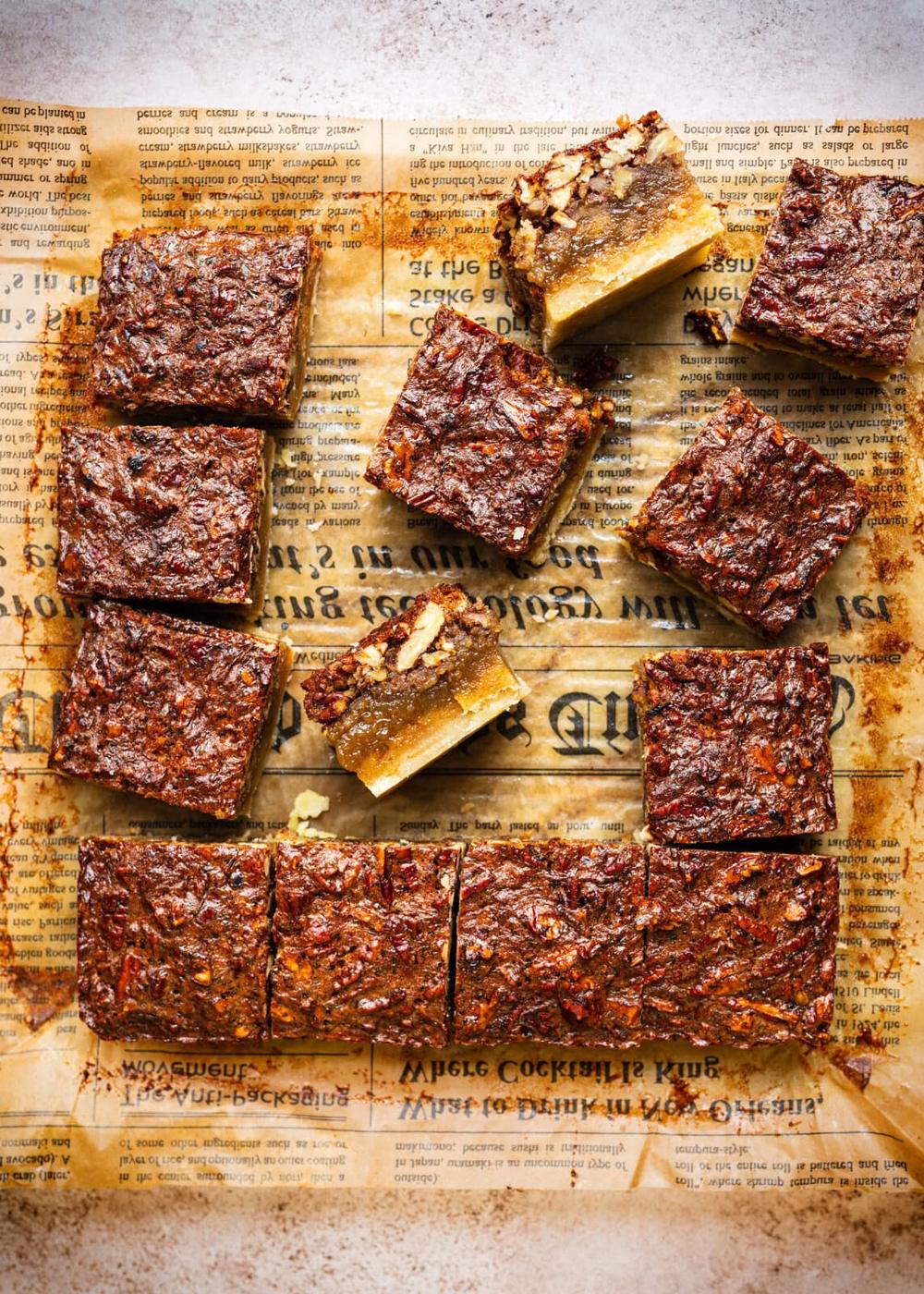 Squares of pecan pie bars arranged on a piece of brown parchment paper printed with text, showing a top view of the rich, nutty dessert with a golden crust and pecan topping.