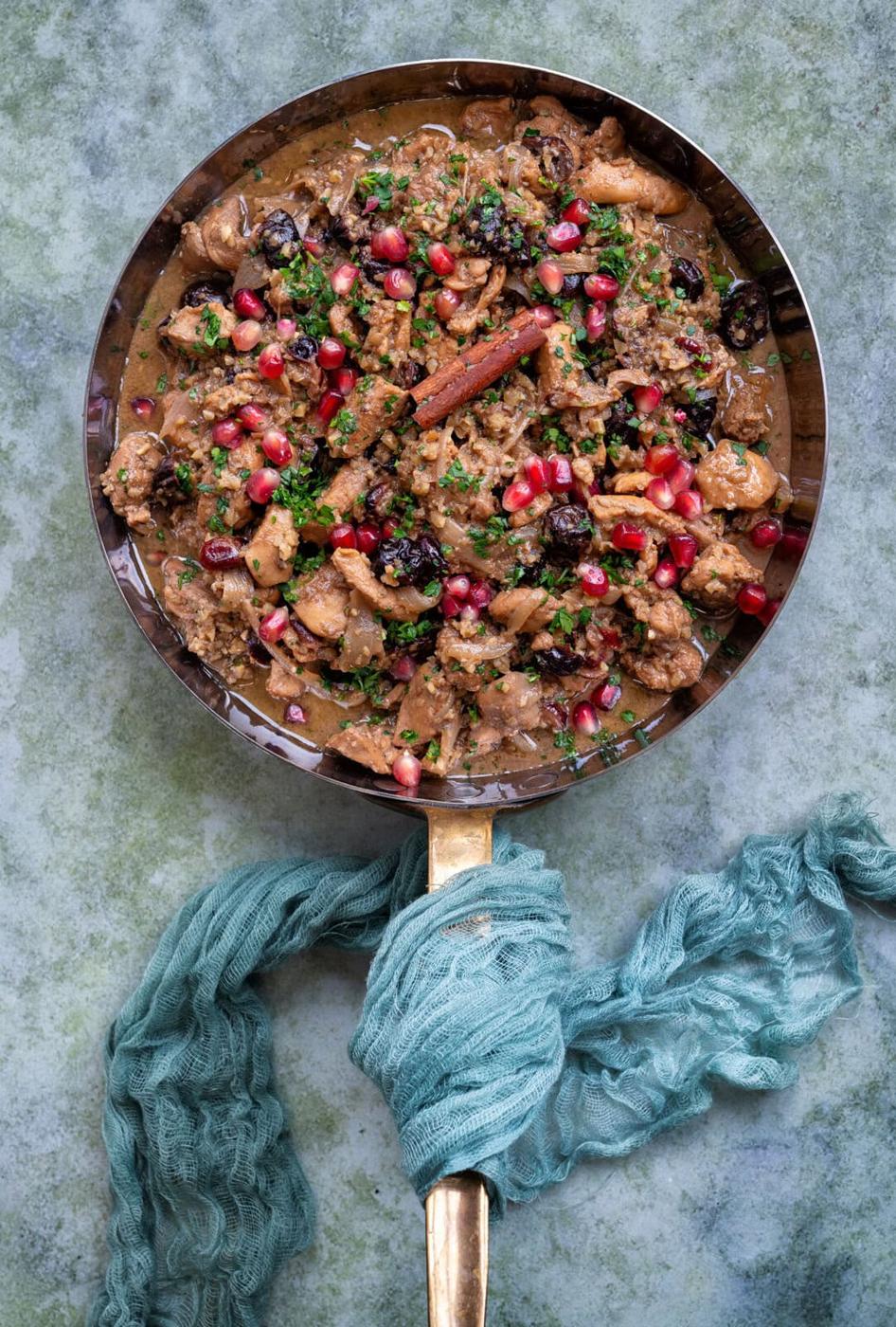 A pan with a golden handle contains a colorful stew of chicken, garnished with pomegranate seeds, herbs, and a cinnamon stick. The pan rests on a textured, green surface, with a teal cloth wrapped around the handle.