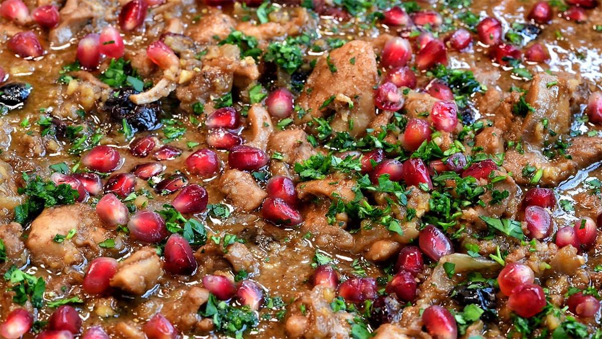 Close-up of a vibrant dish featuring chicken pieces cooked in a rich, brown sauce, generously topped with pomegranate seeds and fresh green herbs, creating a colorful and appetizing presentation.