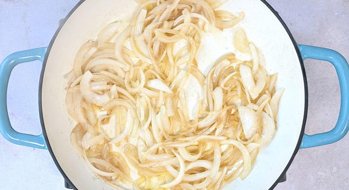 Sliced onions cooking in a white pot with blue handles. The onions are light golden, indicating they are starting to caramelize.