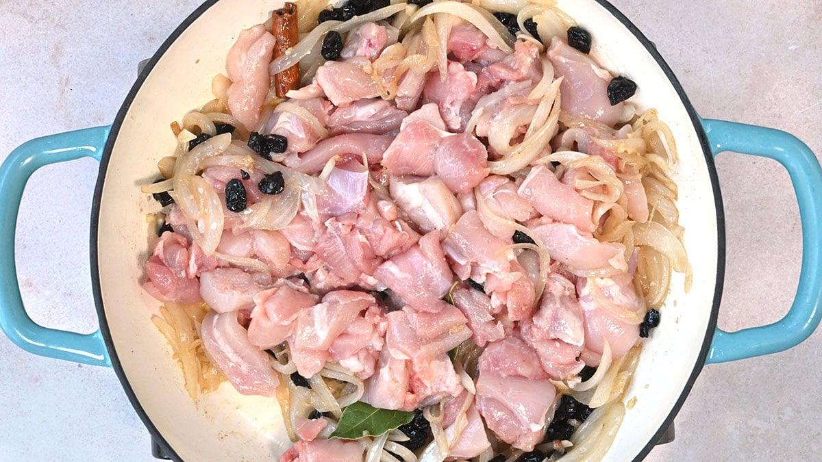 Raw, marinated chicken pieces mixed with sliced onions and small dark berries in a large, round blue-handled pan. The mixture is evenly spread, ready to be cooked.