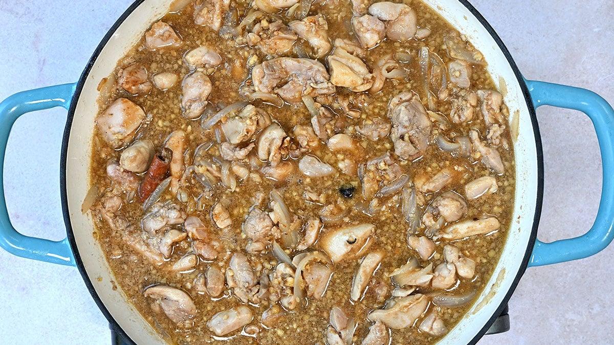 A blue-handled pot filled with a chicken stew containing chunks of chicken, ground walnuts, and visible pieces of onion. The mixture appears to be simmering, with small bubbles on the surface.