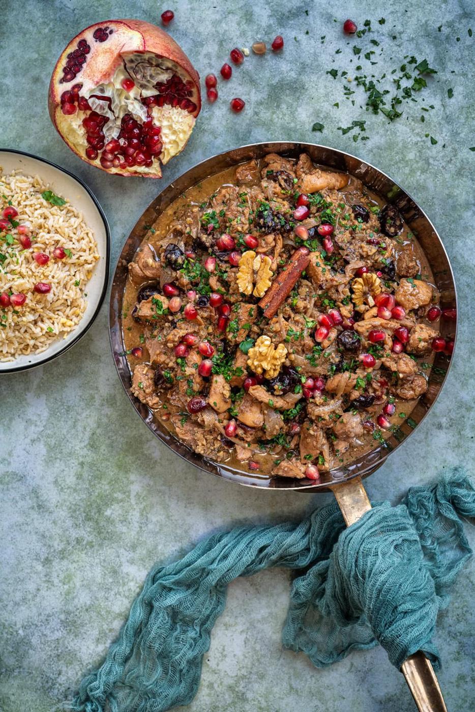 A skillet with Persian stew garnished with walnuts and pomegranate seeds. A bowl of rice topped with herbs and seeds is beside it. A halved pomegranate is also visible. A teal cloth is wrapped around the skillets handle on a textured surface.