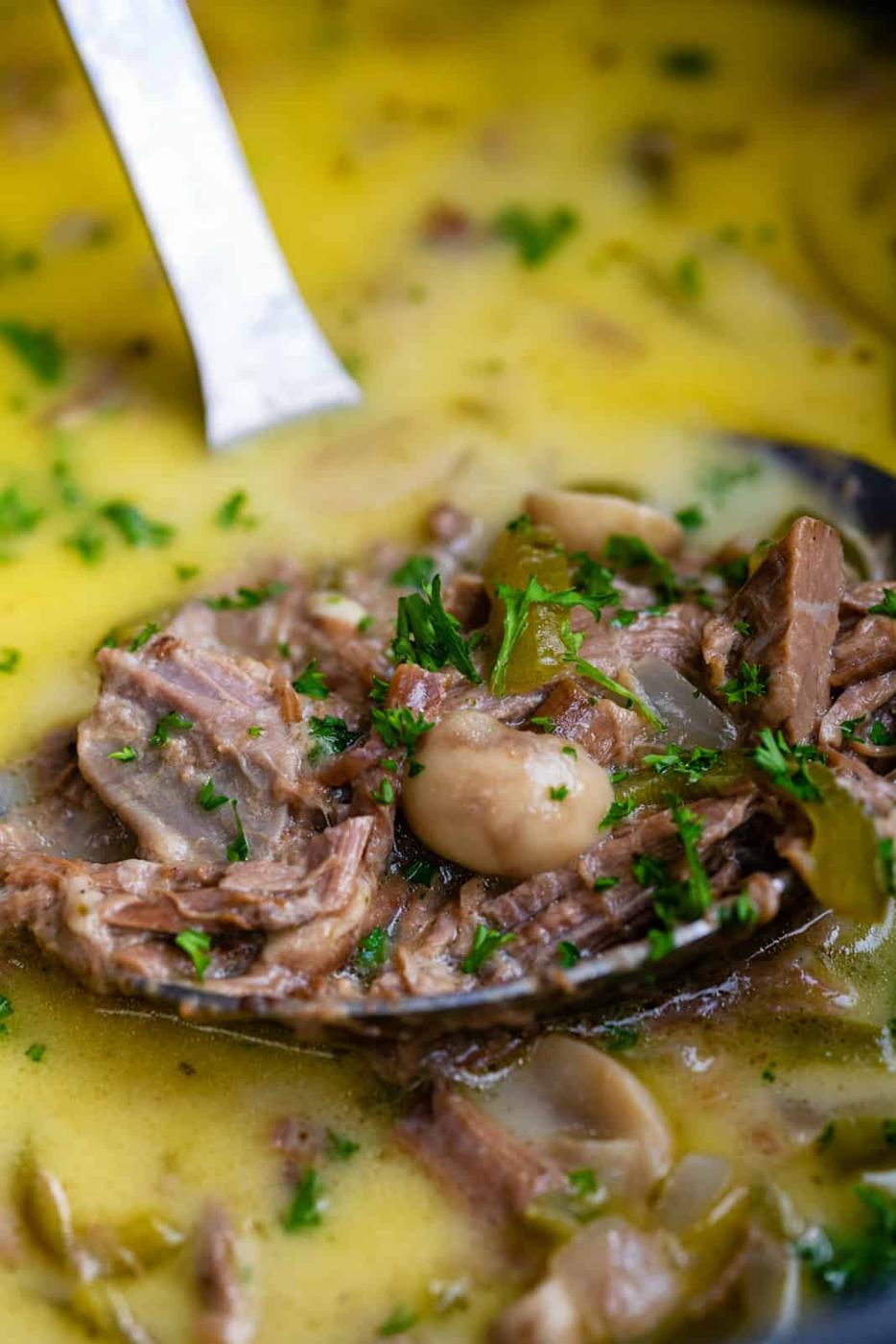 A close-up of a ladle holding shredded beef, mushrooms, and herbs in a creamy yellow broth, garnished with chopped parsley—this Low Carb Philly Cheesesteak Soup is hearty and flavorful.
