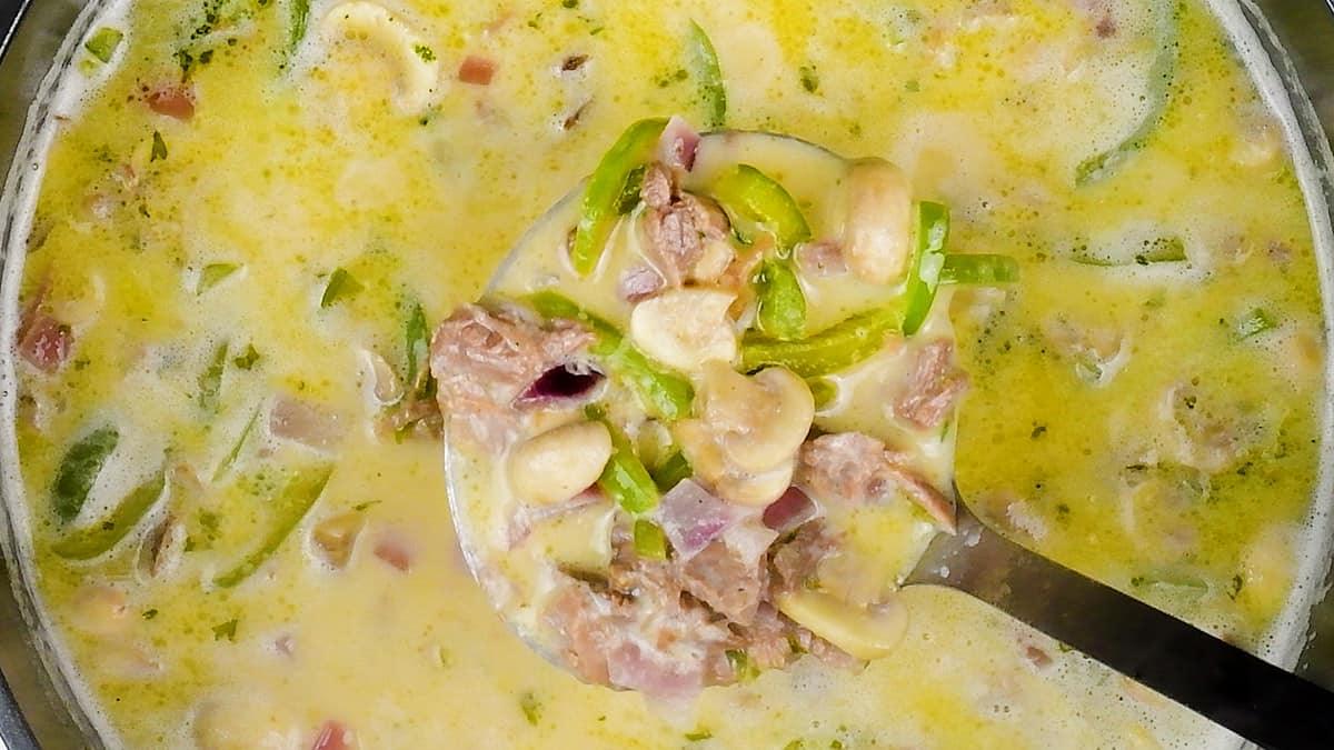 A ladle lifts creamy Keto Philly Cheesesteak Soup, filled with chunks of meat, mushrooms, green peppers, and onions from a pot. The rich, yellowish soup showcases herbs and vibrant vegetables for a hearty meal.