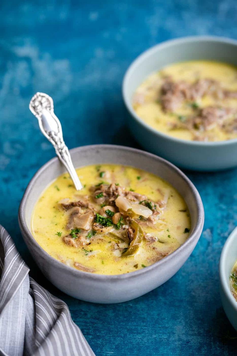 A bowl of creamy Philly Cheesesteak Soup with ground meat, mushrooms, and herbs, served with a spoon. Another bowl is in the background, all set on a blue surface with a striped napkin nearby.