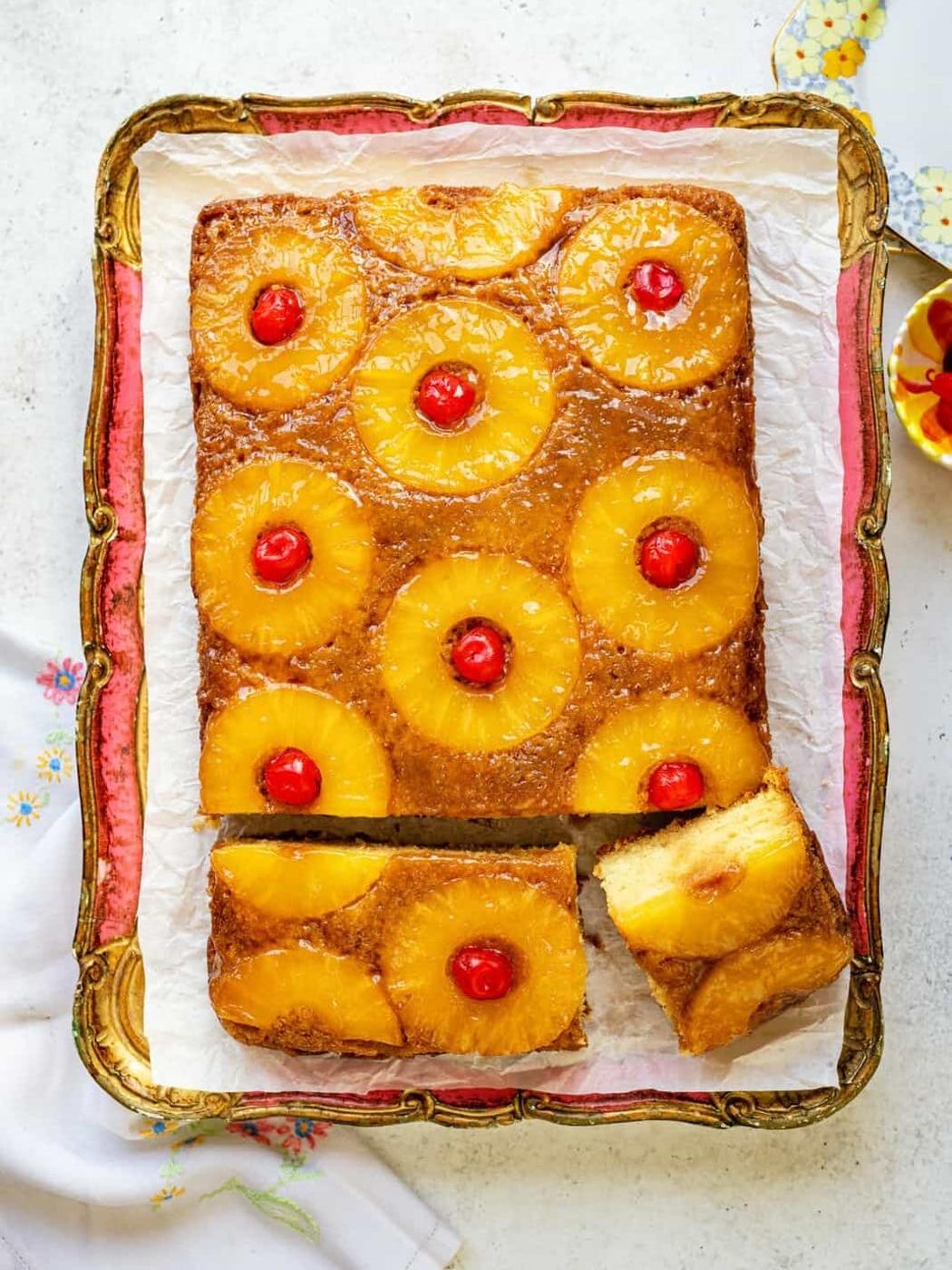 A rectangular Pineapple Upside Down Cake topped with pineapple rings and red cherries sits on a tray. One slice is cut and slightly pulled out, revealing the moist, yellow cake inside.