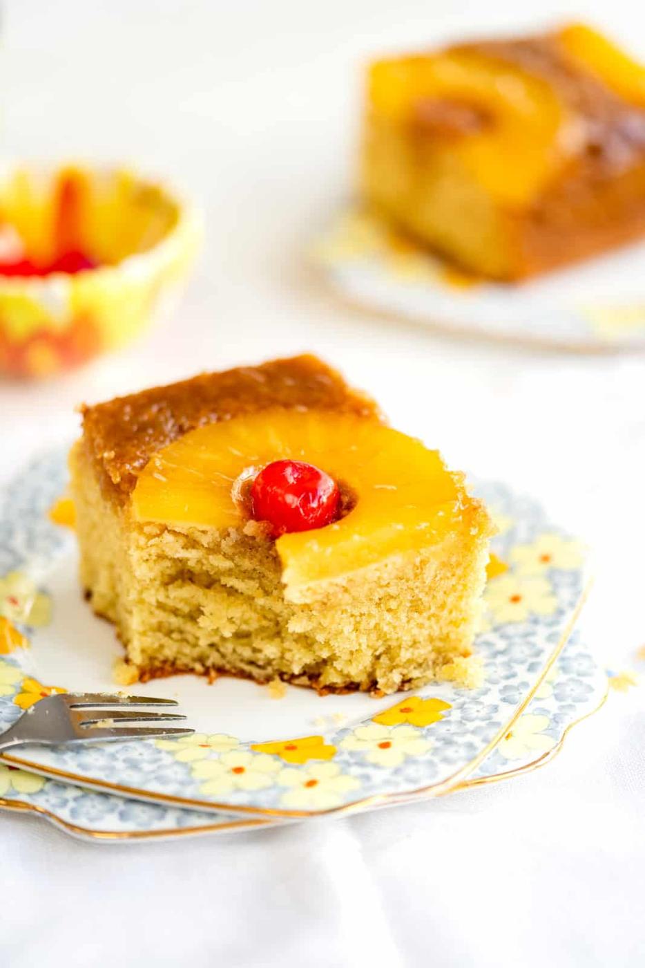 A slice of Pineapple Upside Down Cake with a cherry on top sits on a floral plate, a fork beside it. Another piece of this classic cake is blurred in the background.