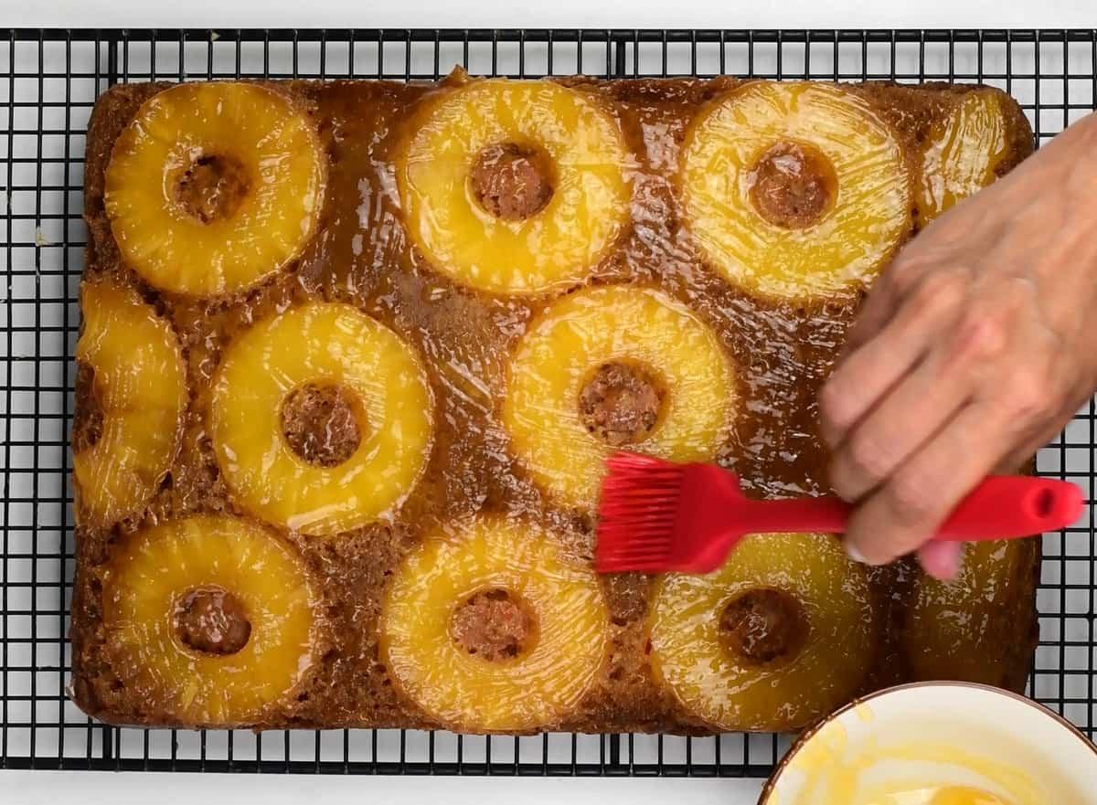 A hand uses a red brush to glaze a Pineapple Upside Down Cake, topped with pineapple rings and cherries, resting on a cooling rack.