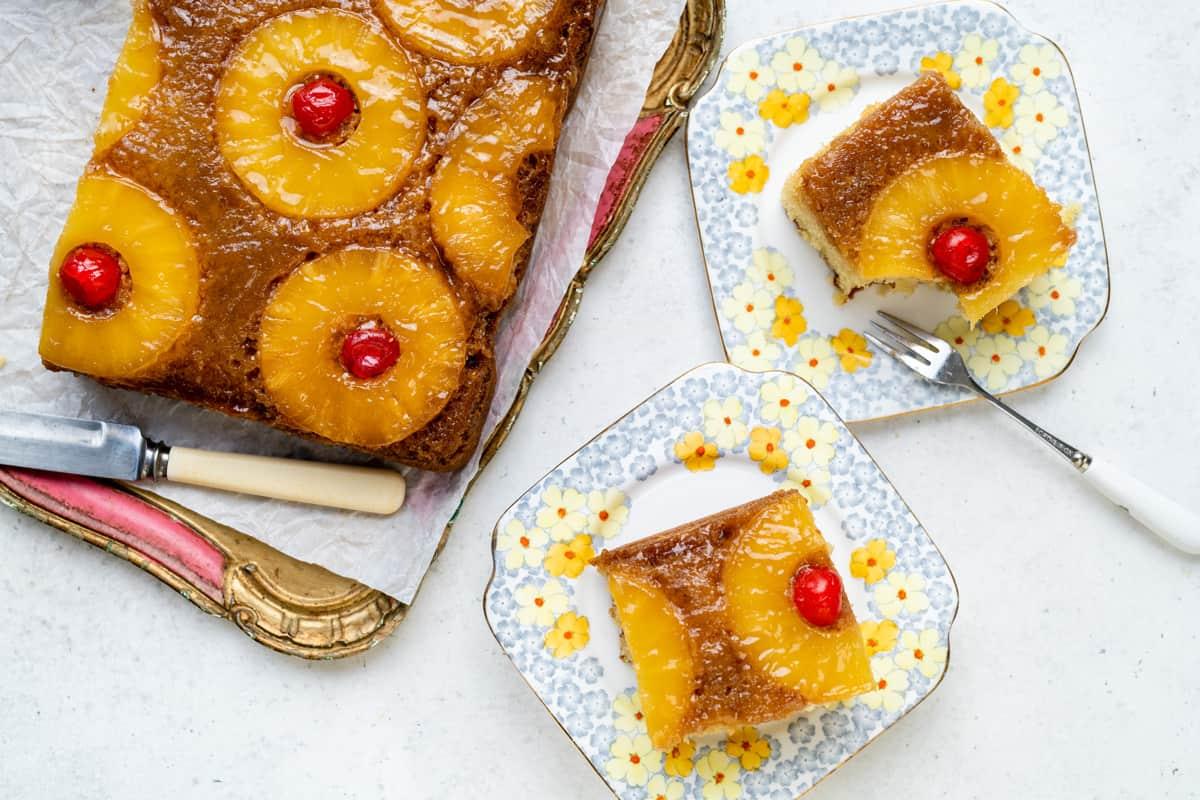 A rectangular Pineapple Upside Down Cake with cherries sits on a tray, with two slices served on floral-patterned plates nearby. A fork rests on one plate, inviting you to enjoy this classic dessert.