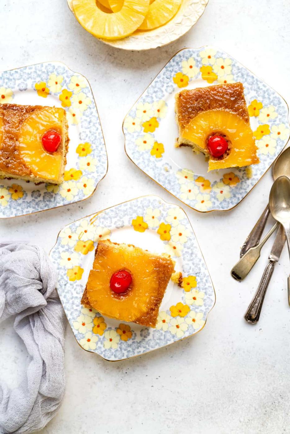 Three slices of Pineapple Upside Down Cake with cherries on top are served on floral-patterned plates. Two forks and a spoon rest nearby, while a dish with extra pineapple rings sits at the top of the image.