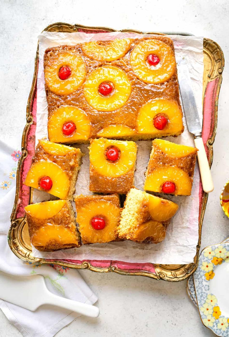 A rectangular Pineapple Upside Down Cake, topped with pineapple rings and maraschino cherries, is sliced into squares on a parchment-lined tray. A serving spatula rests nearby, ready to serve this classic treat.