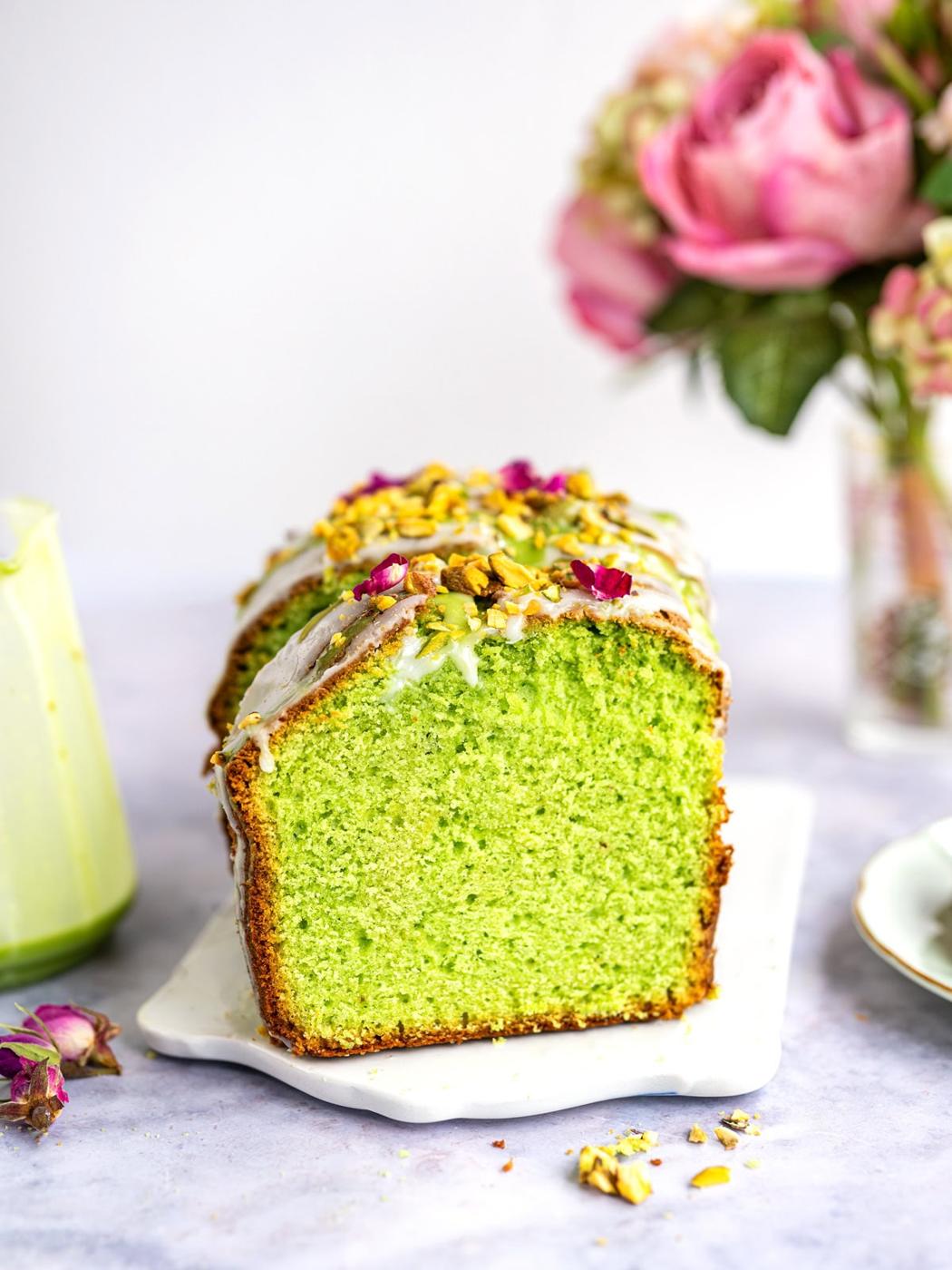 A sliced loaf of bright green cake with white icing and sprinkled pistachios on top sits on a white serving board. In the background, there is a vase of pink flowers and a glass container.