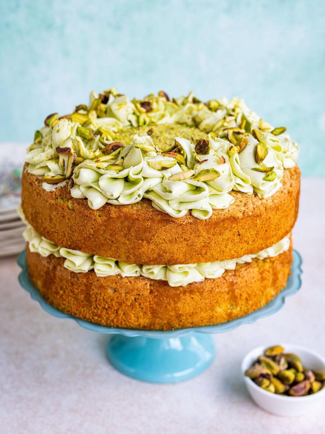A two-layer cake with pale green pistachio frosting and chopped pistachios on top, displayed on a blue cake stand. A small bowl of pistachios sits beside it on a light surface.