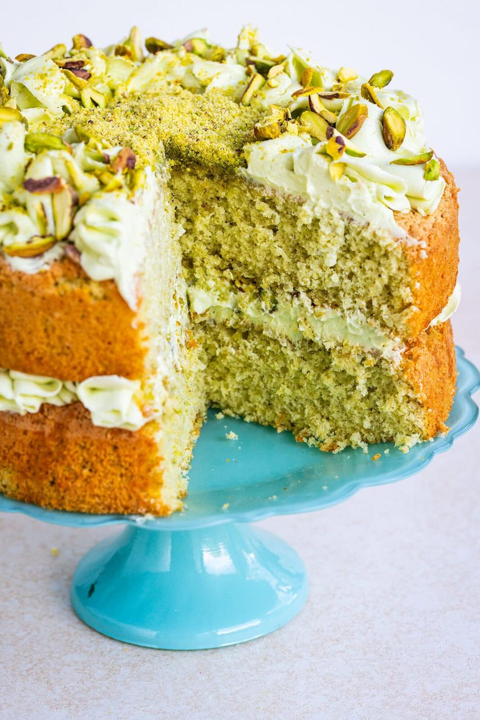 A sliced pistachio cake with green frosting and chopped pistachios on top, displayed on a light blue cake stand.
