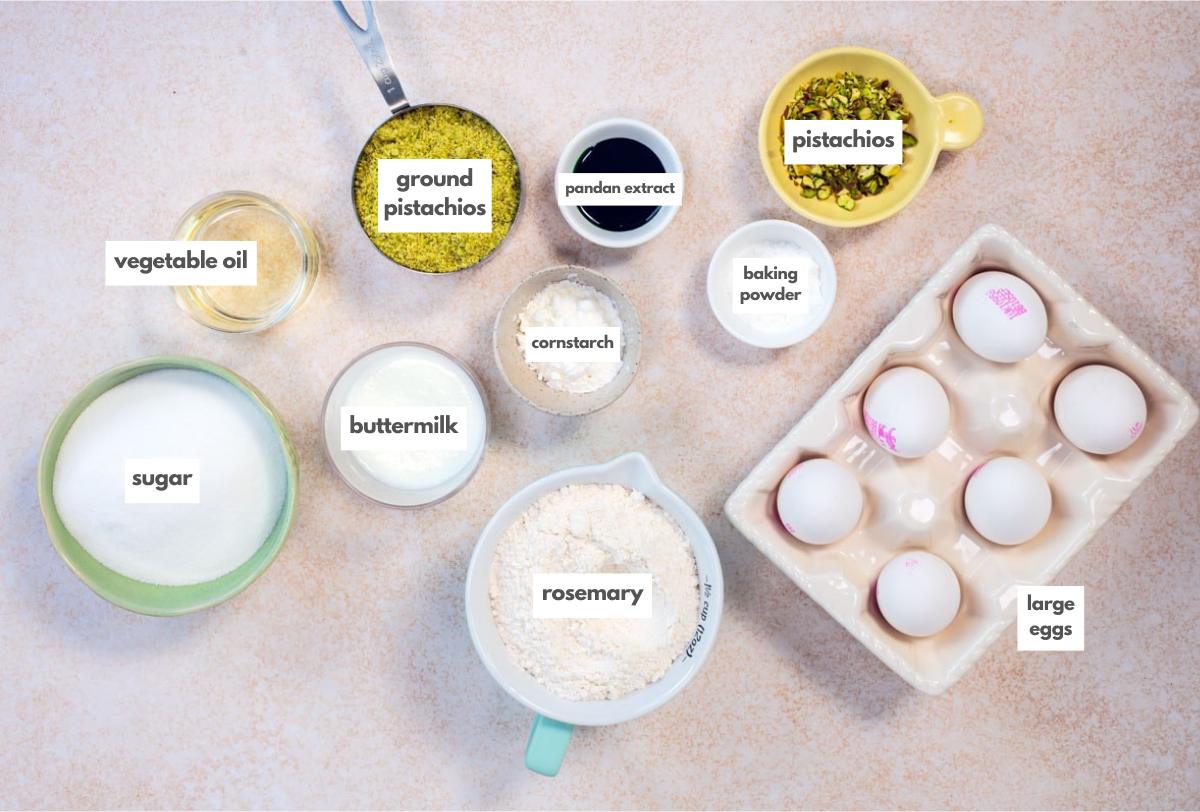 Top-down view of baking ingredients: vegetable oil, sugar, ground pistachios, buttermilk, rosemary, pandan extract, cornstarch, baking powder, chopped pistachios, and six large eggs in a carton. Labels identify each item.