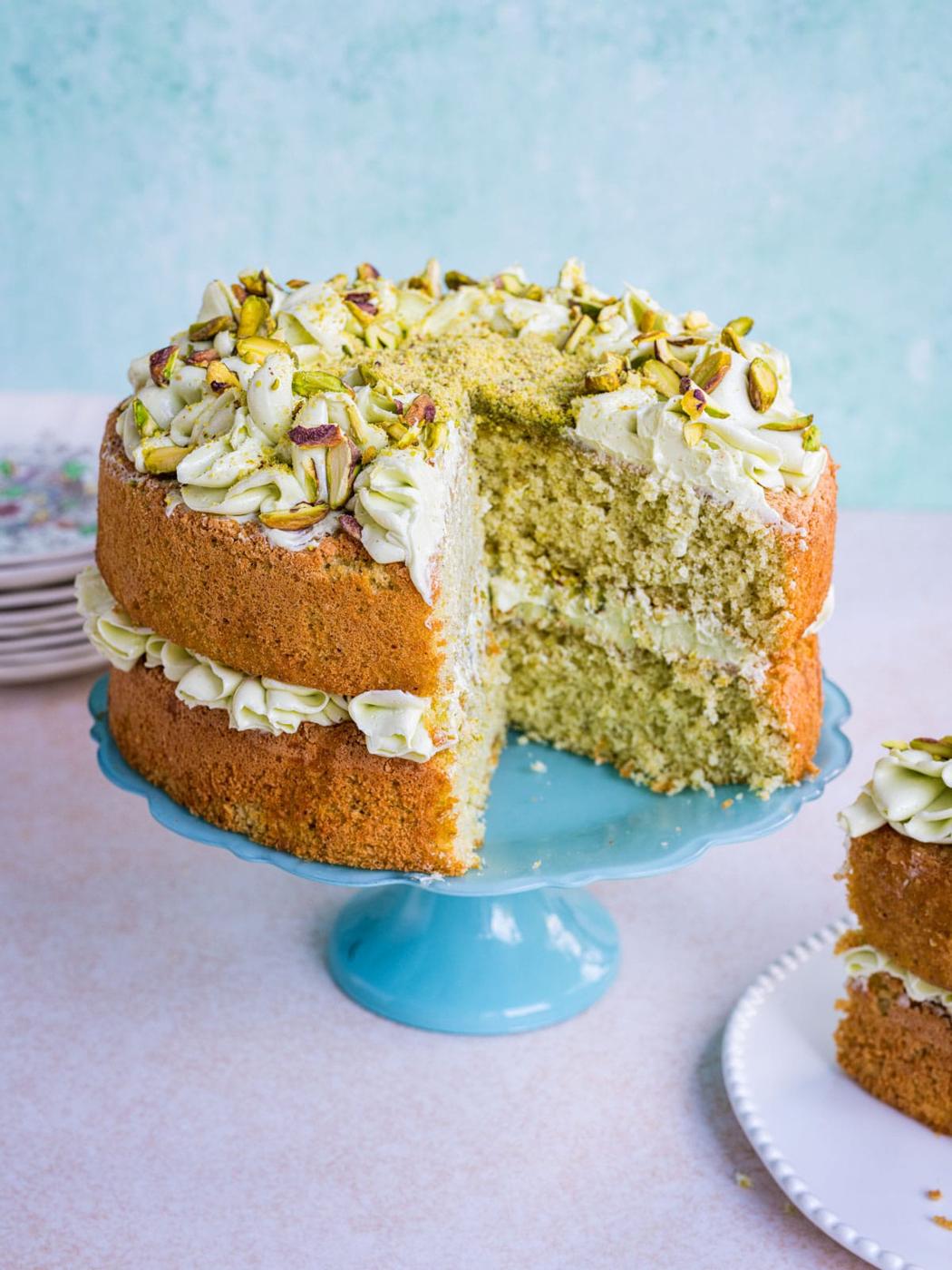 A two-layer pistachio cake with creamy frosting and chopped pistachios on top, displayed on a blue cake stand. A large slice has been cut and placed on a nearby white plate.