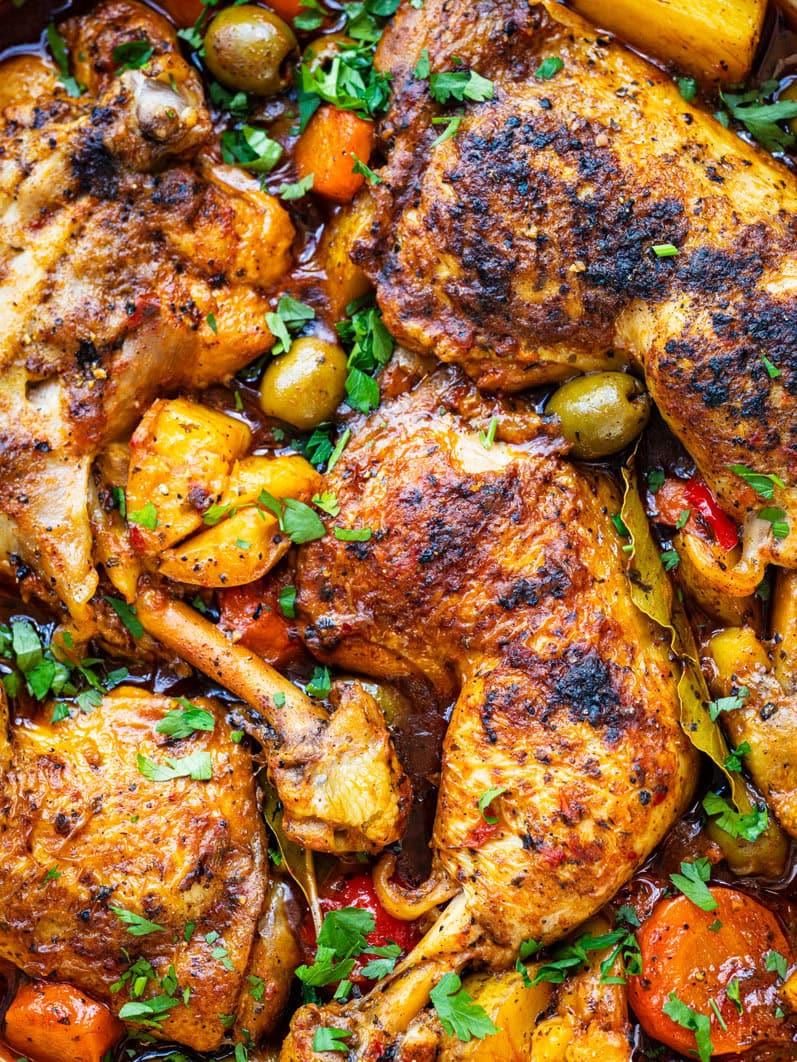 Stewed chicken legs with golden, crispy skin, surrounded by carrots, potatoes, olives, and herbs in a rich, colorful sauce evoke the comforting flavors of pollo guisado, garnished with fresh parsley.