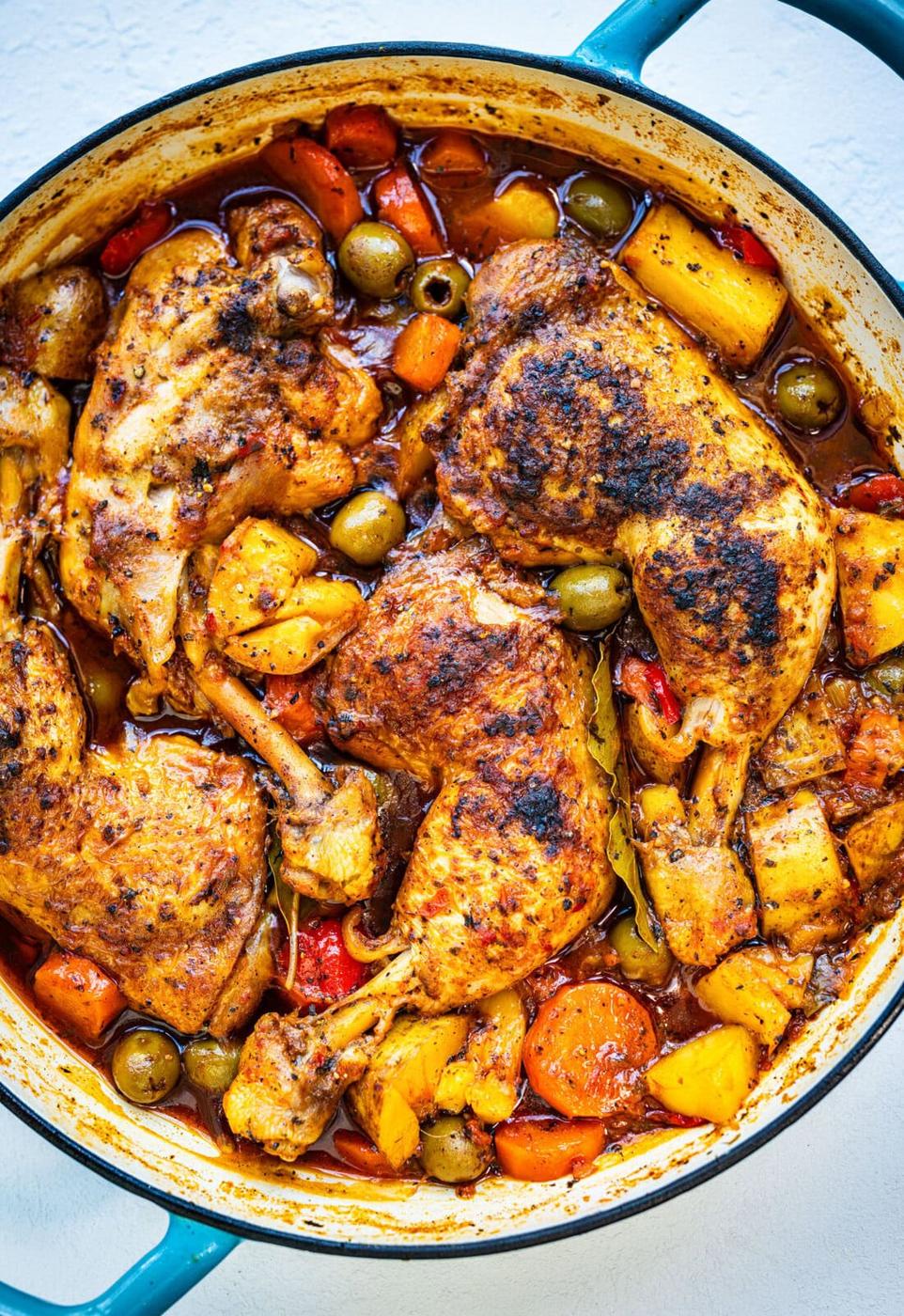 A blue Dutch oven filled with hearty Puerto Rican Pollo Guisado with chicken legs, potatoes, carrots, olives and peppers in a rich, tomato based sauce