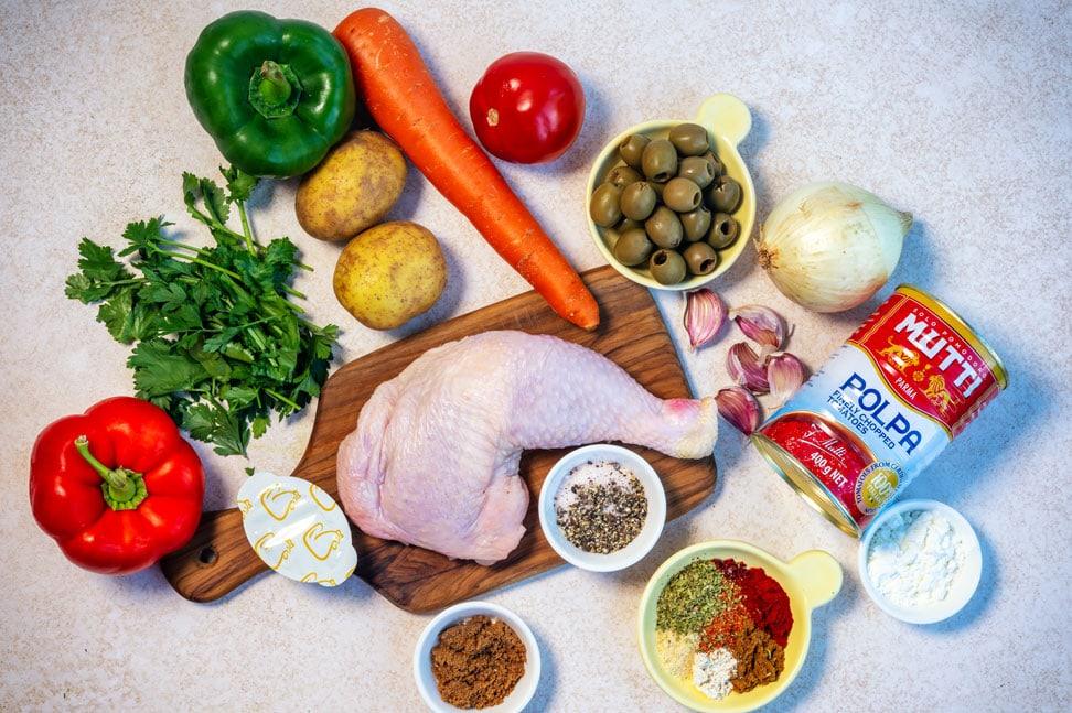 A raw chicken leg on a wooden board surrounded by potatoes, a carrot, parsley, red and green bell peppers, a tomato, onion, garlic, green olives, canned tomatoes, butter, and bowls of various spices on a light surface.