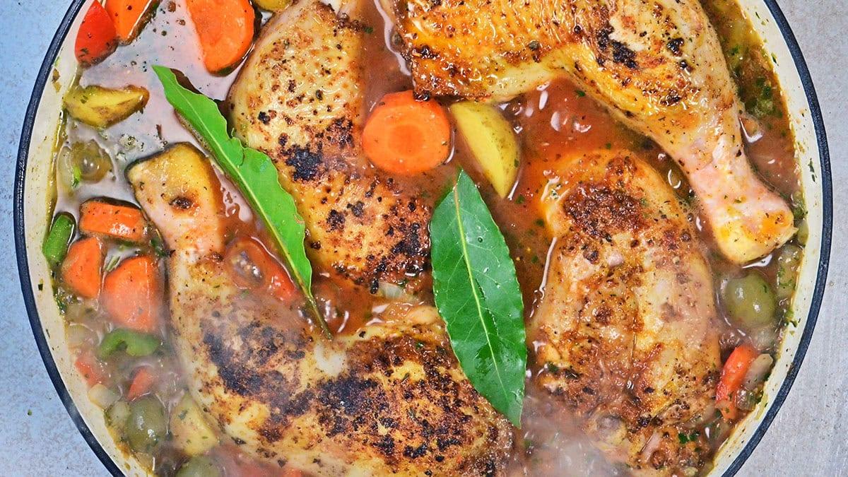 Golden-brown chicken legs simmer in a pot with carrots, potatoes, green olives, and herbs in a rich, savory broth. Two bay leaves rest on top of the stew. Steam rises from the hot dish.