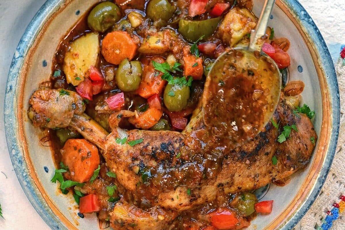 A hand spoons sauce over a baked pollo guisado chicken leg with carrots, potatoes, green olives, and chopped tomatoes in a ceramic dish on a white surface.
