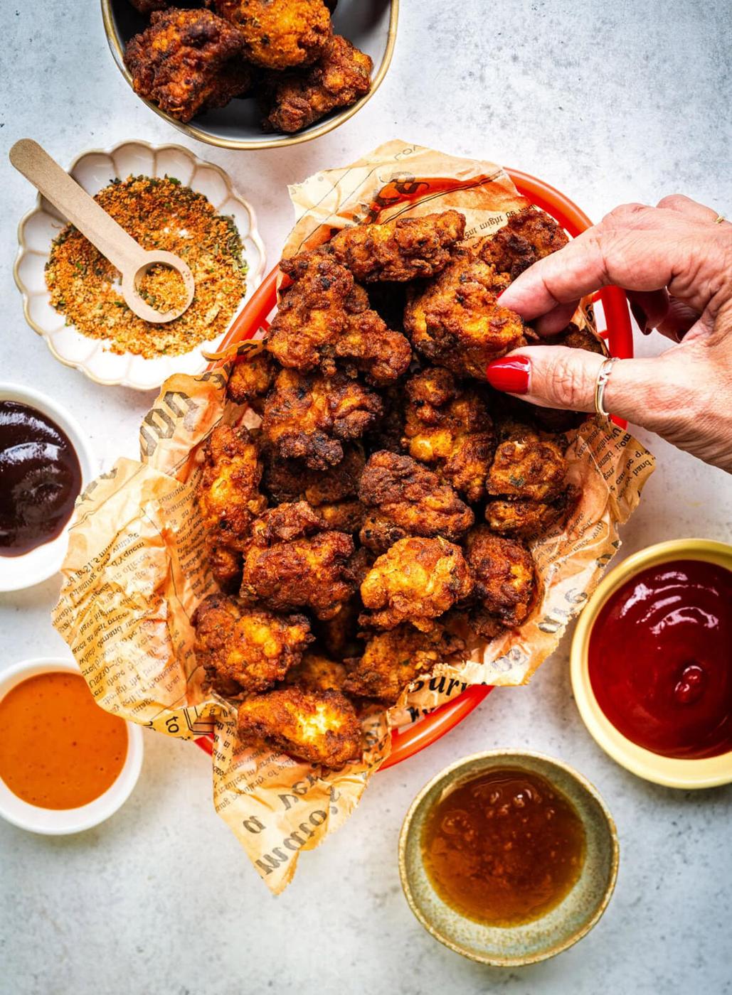 A basket of crispy fried popcorn chicken pieces on newspaper with a hand picking one up, surrounded by bowls of dipping sauces and a small plate of seasoning on a light surface.